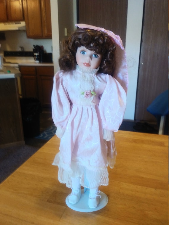 Porcelain Doll # 105 in pink dress with pink bow. Circa. 1980s with metal stand up stand.