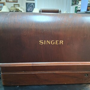 May include: A vintage Singer sewing machine case made of dark wood. The word "SINGER" is displayed in gold lettering on the front. The case has a handle on top and a rectangular shape.
