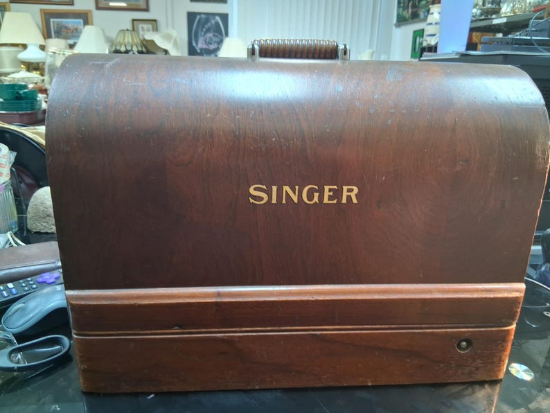May include: A vintage Singer sewing machine case made of dark wood. The word "SINGER" is displayed in gold lettering on the front. The case has a handle on top and a rectangular shape.
