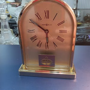 May include: A gold-toned Howard Miller quartz clock with a rectangular base and arched top. The clock face is a light champagne color with black Roman numerals and hands. The clock has a small rectangular emblem at the bottom.