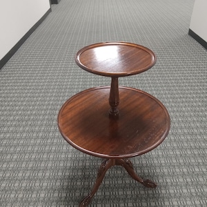 May include: A two-tiered, dark brown wooden side table. The table has a round top and a smaller round tier above it, connected by a central column. The table rests on three curved legs with claw feet. The wood has a glossy finish.