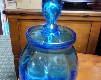 May include: A large, clear blue glass jar with a matching lid and a bulbous body. The lid has a rounded handle. The jar is sitting on a wooden surface. The jar is likely used for storage or decoration.