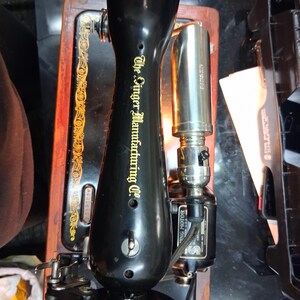 May include: A vintage black Singer Manufacturing Co. sewing machine with gold accents. The machine features a black base, a wooden frame, and a silver-colored light. The text "The Singer Manufacturing Co." is visible on the machine.