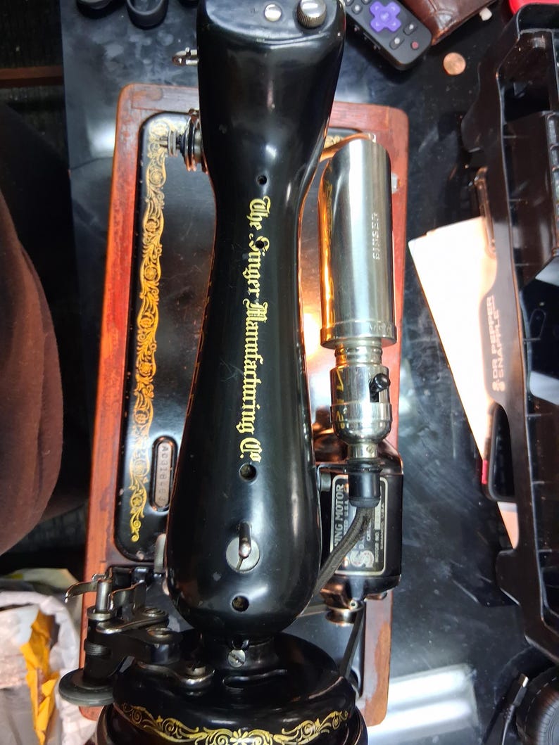 May include: A vintage black Singer Manufacturing Co. sewing machine with gold accents. The machine features a black base, a wooden frame, and a silver-colored light. The text "The Singer Manufacturing Co." is visible on the machine.