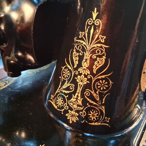 May include: Close-up of a vintage black sewing machine with gold floral detailing. The machine's body is black with intricate gold leaf designs. The gold patterns include flowers, leaves, and swirling lines, adding a decorative touch to the machine.
