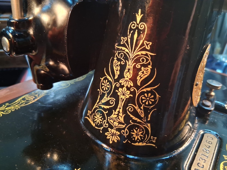 May include: Close-up of a vintage black sewing machine with gold floral detailing. The machine's body is black with intricate gold leaf designs. The gold patterns include flowers, leaves, and swirling lines, adding a decorative touch to the machine.