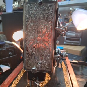 May include: Close-up of a vintage sewing machine. The machine features ornate floral engravings on a silver-colored panel. The machine has a black base with gold accents and a needle in the center.