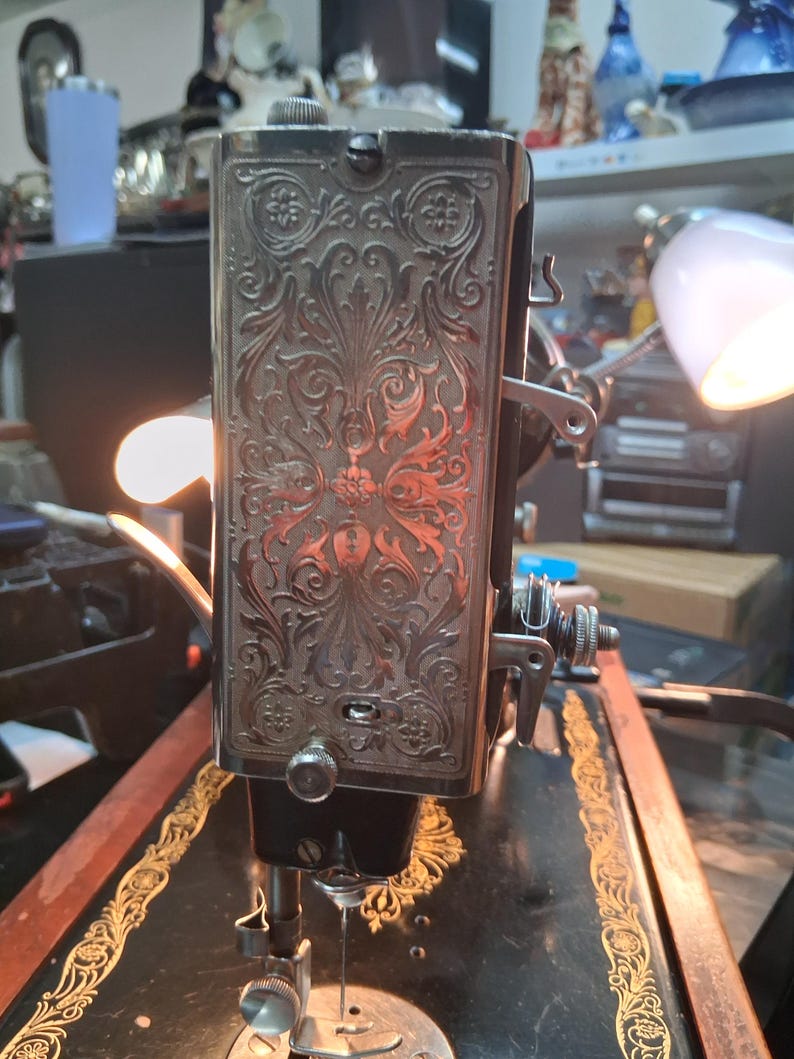 May include: Close-up of a vintage sewing machine. The machine features ornate floral engravings on a silver-colored panel. The machine has a black base with gold accents and a needle in the center.