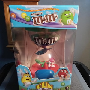 May include: A red, blue, and green plastic candy dispenser shaped like an hourglass with a spinning wheel at the bottom. The dispenser is packaged in a clear plastic box with a colorful cardboard box surrounding it. The box features the M&M's logo and the text "Fun Candy Dispenser Machine!" and "9.40 oz bag of M&M's Brand Chocolate Candies Included".
