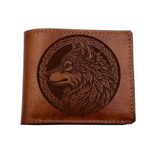 May include: Brown leather wallet featuring a detailed wolf design within a circular frame. The wolf is intricately etched, showcasing fur and facial features. The wallet has a classic bi-fold design with visible stitching.