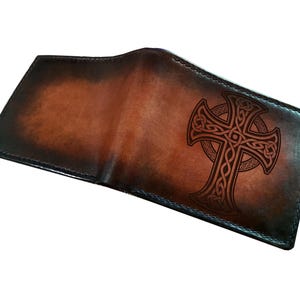 May include: Open brown leather wallet featuring a Celtic cross design. The wallet has a gradient effect, transitioning from dark to lighter brown shades. The cross is intricately detailed.