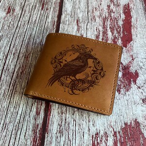 May include: A tan leather wallet with a detailed illustration of a raven inside a circular frame. The wallet is rectangular, with visible stitching around the edges. The raven design is in a darker shade of brown.