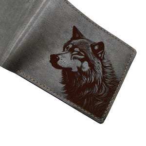 Puede incluir: Cartera de cuero gris con un retrato detallado de un lobo grabado en la parte delantera. La cartera está abierta, revelando el interior. Los bordes están cosidos con un hilo en contraste, y el diseño es minimalista y robusto.