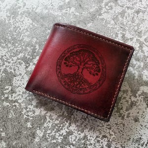 Könnte beinhalten: Eine bordeauxfarbene Lederbrieftasche mit einem Lebensbaum-Design. Die Brieftasche ist rechteckig und weist eine detaillierte kreisförmige Gravur eines Baumes mit Wurzeln auf. Die Nähte sind an den Rändern sichtbar.