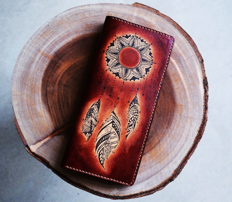 May include: A brown leather wallet with a mandala design and a dreamcatcher motif. The wallet has a flap closure and is decorated with intricate details.