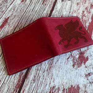 Può includere: Un portafoglio bifold rosso scuro con un'illustrazione scura e dettagliata incisa al laser di un grifone sul pannello frontale. Il portafoglio presenta cuciture tonali visibili lungo i bordi e una finitura opaca e liscia sulla superficie.