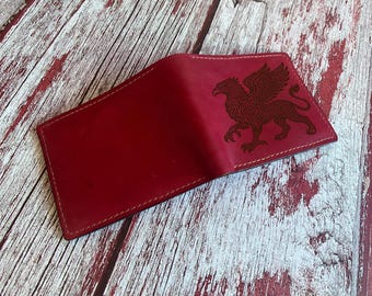 May include: A deep red bifold wallet featuring a dark, intricate laser-engraved illustration of a griffin on the front panel. The wallet is constructed with visible tonal stitching along the edges and has a smooth, matte finish on the surface.