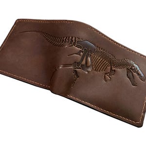 May include: Brown leather wallet with a detailed embossed image of a dinosaur skeleton. The wallet is open, revealing the interior, and features stitched edges. A unique accessory for anyone interested in paleontology.