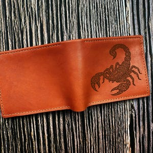 May include: A tan leather bifold wallet featuring a dark brown laser-engraved scorpion design on the front panel. The wallet has visible stitching along the edges and a smooth, matte finish, showcasing a detailed, intricate pattern on the scorpion.
