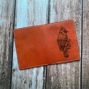 May include: A rectangular tan leather cover featuring a detailed black laser-engraved illustration of a crested bird perched on a floral branch. The leather has visible stitching along the edges and a smooth, matte finish across the surface.