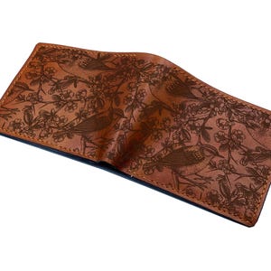 May include: A brown leather bifold wallet featuring a detailed laser-engraved pattern of small birds perched on flowering branches. The wallet has visible stitching along the edges and a smooth, matte finish across the exterior surface.