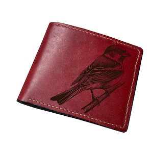 May include: A red leather wallet with a detailed engraving of a bird perched on a branch. The wallet is rectangular, with visible stitching around the edges. The bird design is in a darker shade, creating a contrast.