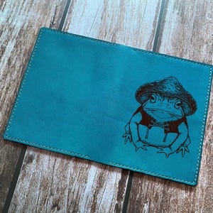 May include: Teal leather notebook cover with a frog illustration wearing a mushroom hat and vest. The frog is in dark brown. The cover has white stitching around the edges. The notebook cover is a rectangular shape.