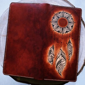 May include: A brown leather journal with a black and white dreamcatcher design. The dreamcatcher features a mandala-like sun with feathers hanging from it.