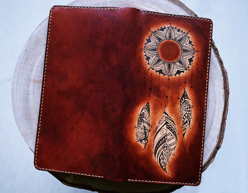 May include: A brown leather journal with a black and white dreamcatcher design. The dreamcatcher features a mandala-like sun with feathers hanging from it.