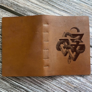 Könnte beinhalten: Ein braunes Lederjournal mit einem genähten Rücken und einem lasergravierten Design einer Schlange in einem geometrischen Muster.