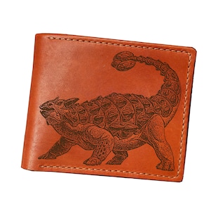 May include: A tan leather wallet with a detailed engraving of an Ankylosaurus dinosaur. The wallet has a bi-fold design with visible stitching around the edges. The dinosaur image is in a darker brown, creating a contrast against the lighter leather.
