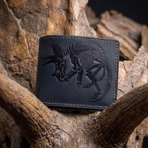 May include: A black leather bifold wallet featuring a debossed skeletal illustration of a triceratops dinosaur on the front cover. The wallet is constructed with visible brown stitching along the edges, creating a contrast against the dark leather.