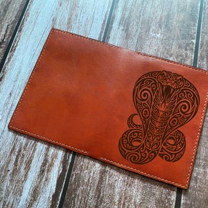 Peut inclure: Une couverture rectangulaire en cuir beige ornée d'un motif de serpent cobra gravé au laser en brun foncé avec des motifs tribaux complexes. Le cuir présente des coutures visibles sur les bords et une surface lisse et plate.