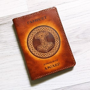 Puede incluir: Cartera de pasaporte de cuero marrón con un grabado detallado de un martillo y un círculo de nudos. El texto "PASSPORT" y "KINGDOM OF ASGARD" está grabado en la cubierta.