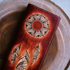 May include: A brown leather wallet with a black and white dreamcatcher design. The dreamcatcher features a mandala-like sun with feathers hanging from it.