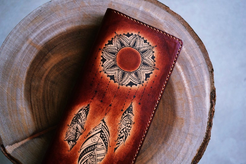 May include: A brown leather wallet with a black and white dreamcatcher design. The dreamcatcher features a mandala-like sun with feathers hanging from it.