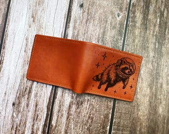 May include: A tan leather bifold wallet featuring a dark brown laser-engraved illustration of a raccoon wearing a space helmet, surrounded by small star shapes. The wallet has visible stitching along the top and side edges.
