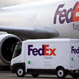 May include: A white FedEx delivery truck with the FedEx Express logo in purple and orange is parked in front of a FedEx airplane. The airplane is white with purple accents and the FedEx logo. The image conveys a theme of shipping and logistics.