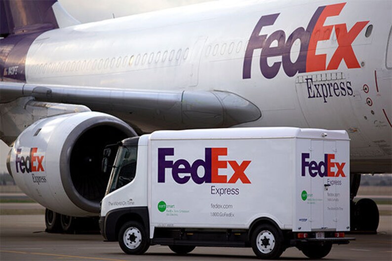 May include: A white FedEx delivery truck with the FedEx Express logo in purple and orange is parked in front of a FedEx airplane. The airplane is white with purple accents and the FedEx logo. The image conveys a theme of shipping and logistics.