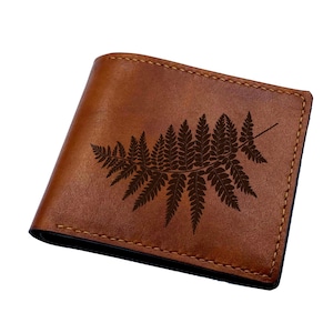 Puede incluir: Cartera de cuero marrón con un diseño de hoja de helecho. La cartera es rectangular con esquinas redondeadas y bordes cosidos. El helecho está grabado en el cuero.