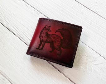 May include: A bifold wallet in a deep gradient of red and black, featuring a detailed, dark-toned engraving of a fox on the front cover. The wallet has visible stitching along the edges and a smooth, polished finish across the leather surface.