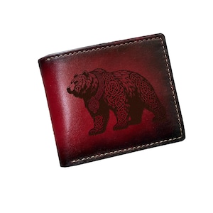 May include: A bi-fold leather wallet with a gradient red and brown finish. The wallet features a detailed bear design with Celtic knot patterns. The stitching is white, and the wallet is rectangular.