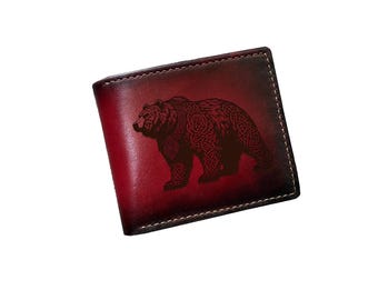May include: A bi-fold leather wallet with a gradient red and brown finish. The wallet features a detailed bear design with Celtic knot patterns. The stitching is white, and the wallet is rectangular.