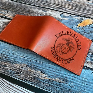 Peut inclure: Un portefeuille en cuir orange ouvert, avec l'emblème et le texte des United States Marine Corps. L'emblème représente un aigle, un globe et une ancre dans un cercle. Le portefeuille est sur une surface en bois vieilli.