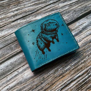 May include: A teal leather wallet featuring a capybara in an astronaut helmet, with stars and lines etched around it. The wallet is closed and has a stitched border. The background is a wooden surface.