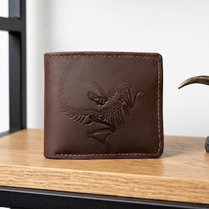 Könnte beinhalten: Eine braune Ledergeldbörse mit einer genähten Kante. Die Vorderseite zeigt ein geprägtes Dinosaurierskelett-Design. Die Geldbörse ist rechteckig und liegt auf einem Holzregal. Rechts befindet sich ein dekoratives Objekt.