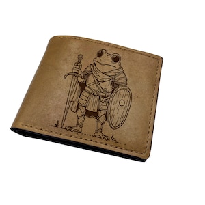 May include: A tan bi-fold wallet featuring a dark brown laser-engraved illustration of a frog dressed in medieval armour, holding a long sword in one hand and a round shield in the other. The design is centred on the front exterior surface.