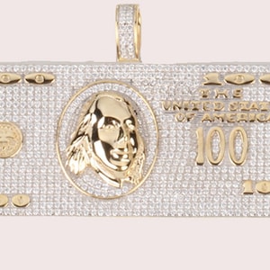 May include: A gold and diamond-encrusted pendant shaped like a $100 bill. The pendant features a portrait of Benjamin Franklin, the words "THE UNITED STATES OF AMERICA", and the number "100" repeated several times.