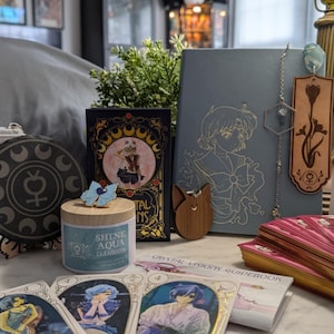 May include: A collection of items including a white canvas pouch with a blue mercury symbol, a circular black moon phase coaster, a candle labeled Shine Aqua Illusion, a light blue notebook with an anime character, a wooden bookmark, and tarot cards.