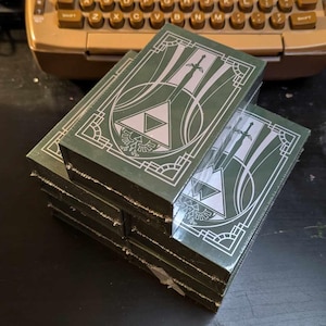 May include: A stack of green cardboard boxes with a white design featuring a stylized triangle and swords. The boxes have a decorative border.